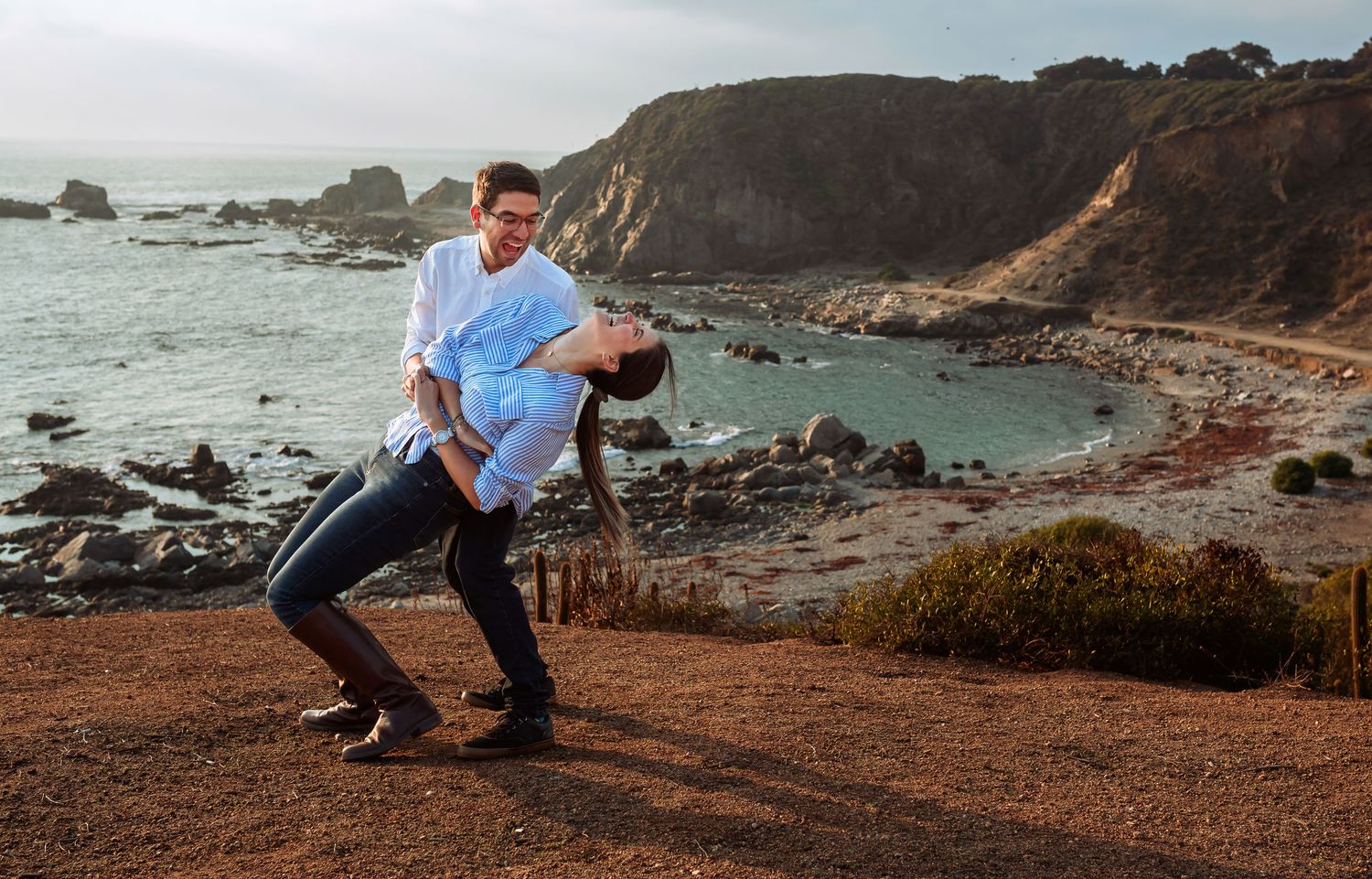Couple dancing romantically on coastal cliffside with scenic ocean cove backdrop.