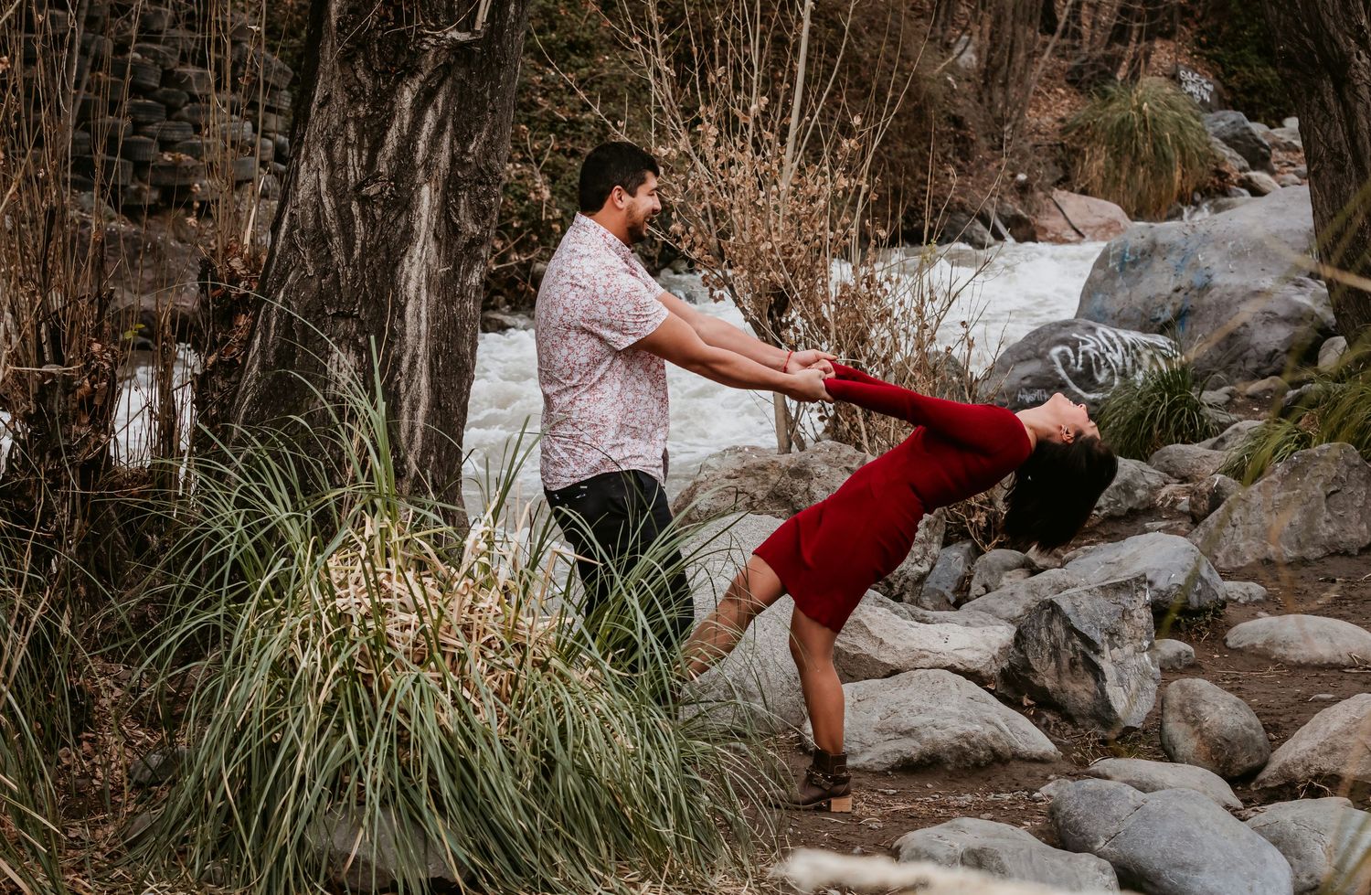 Couple dancing outdoors as man dips woman in red dress among rocks and tall grasses by stream.