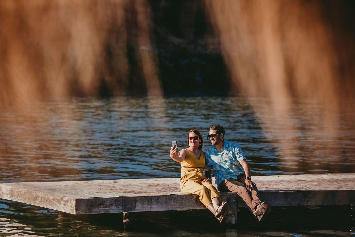 Happy couple sitting on a wooden dock over a lake taking a selfie on a sunny day.