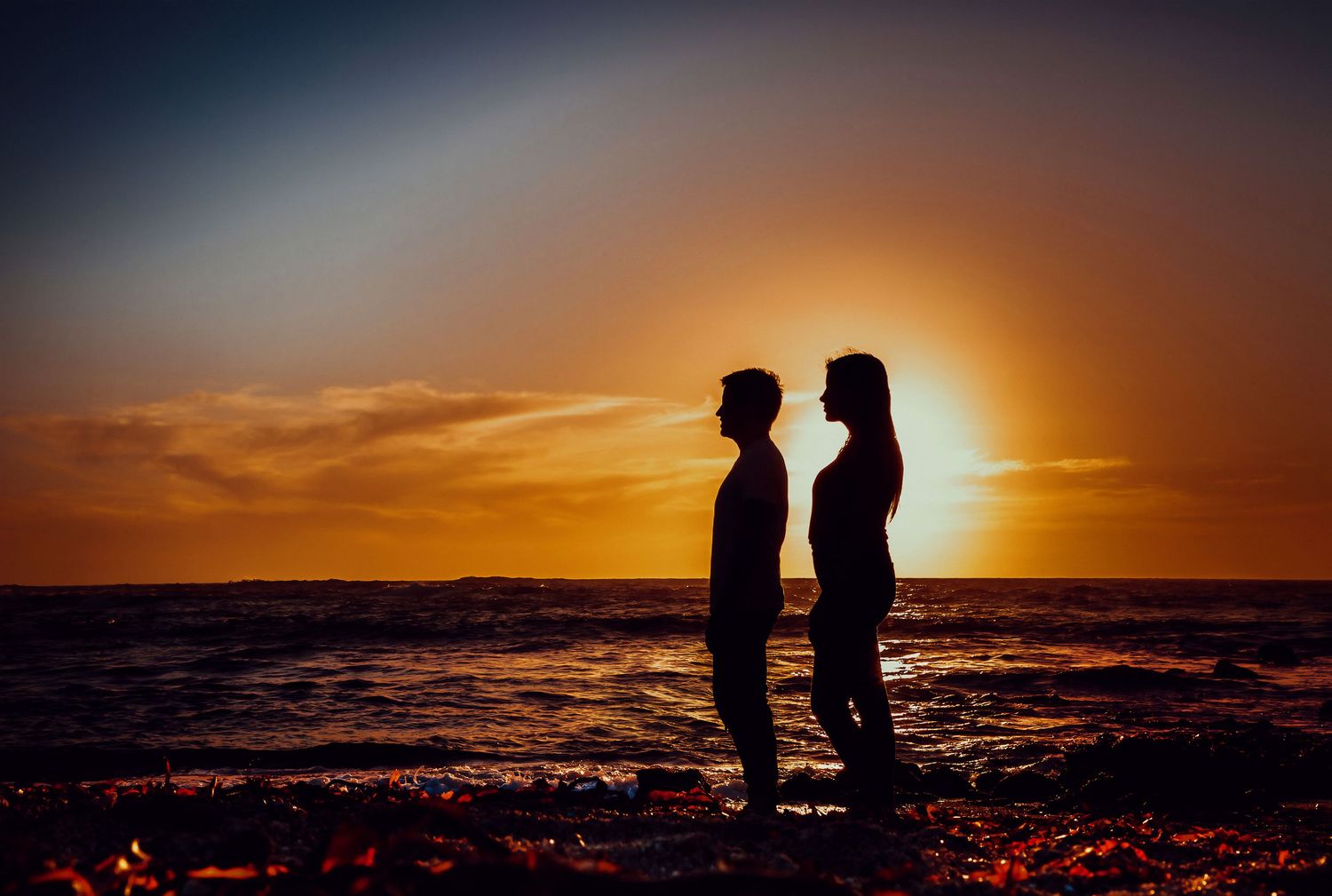 Silhouette of couple facing each other against a vibrant orange ocean sunset.
