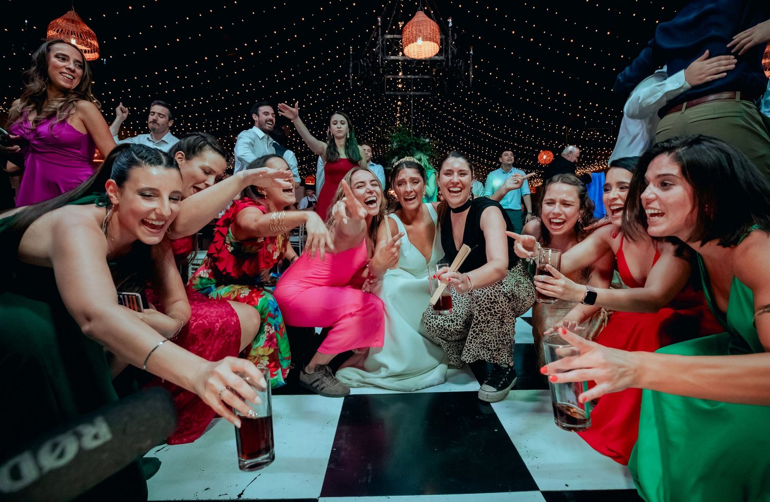 Bride poses for selfie surrounded by cheerful bridesmaids in colorful dresses at wedding reception.