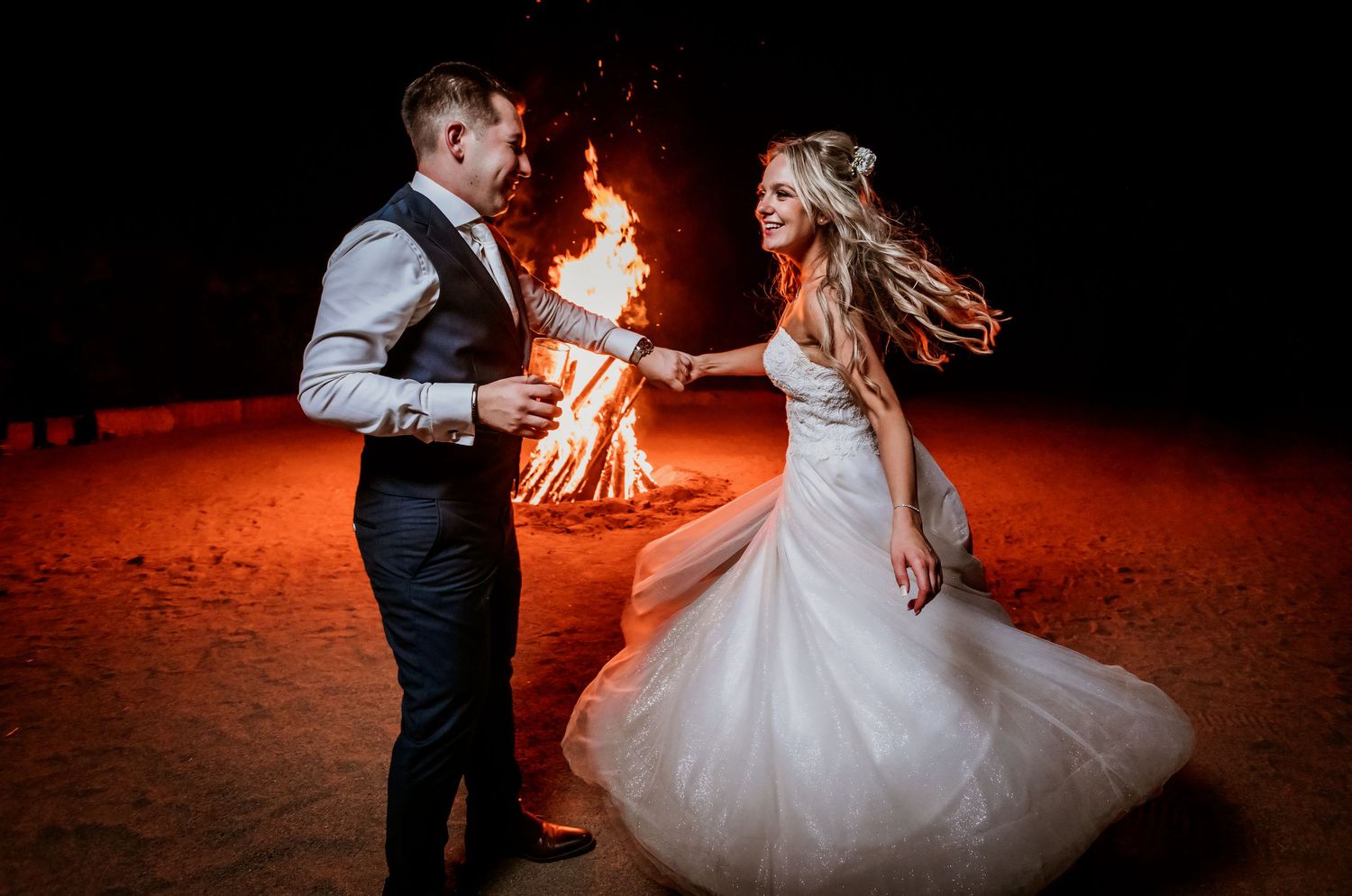 Bride and groom dancing joyfully beside a large bonfire on a sandy beach at night during their wedding.