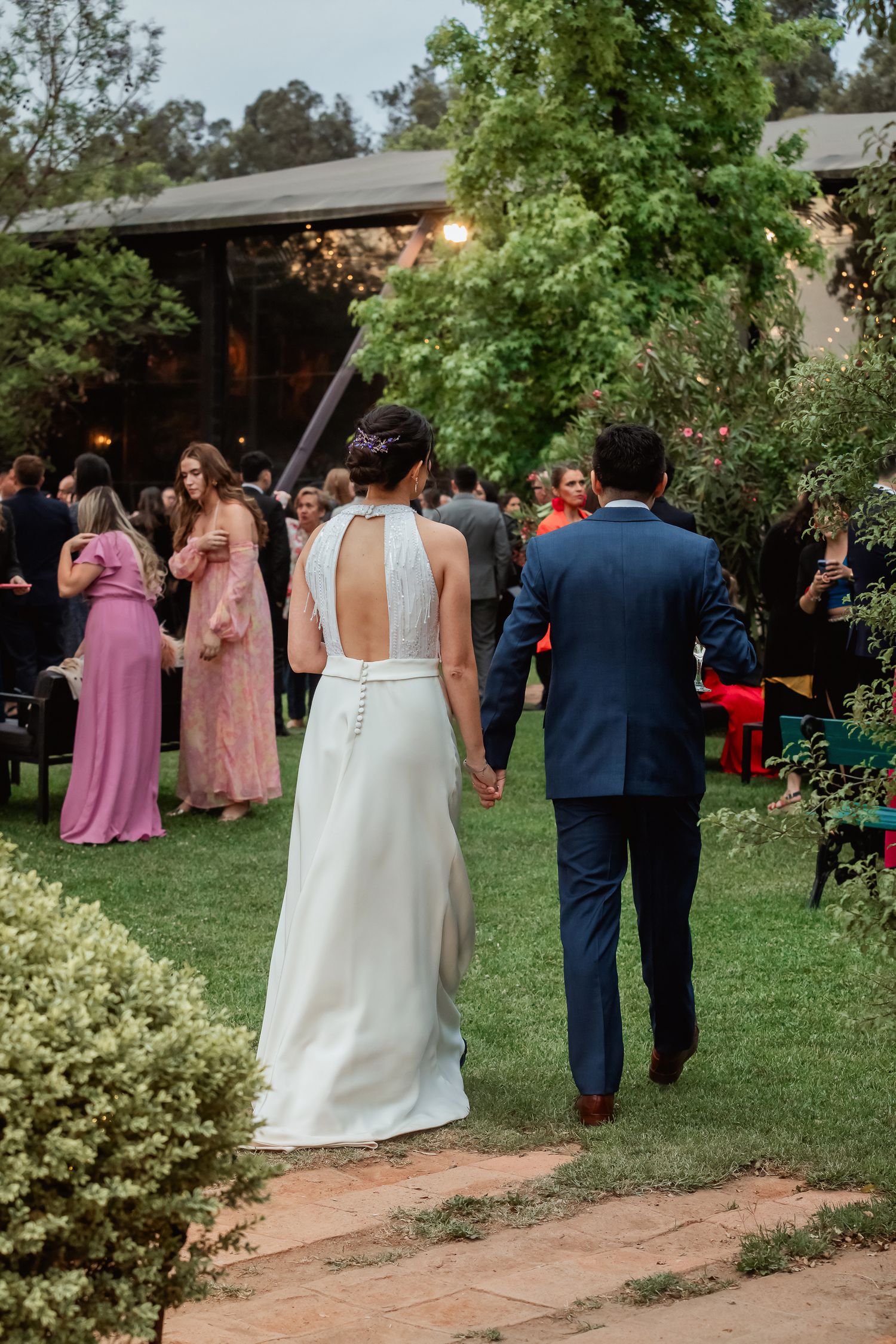 Bride and groom walking hand in hand toward wedding reception guests in an outdoor garden venue.