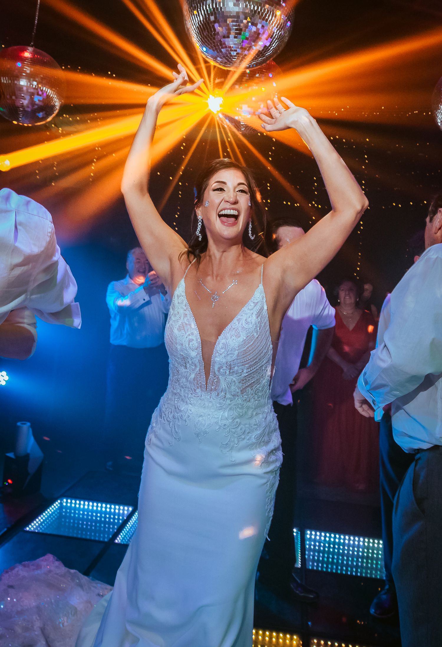 Bride in white gown joyfully raises disco ball overhead on dance floor with colorful lights at wedding reception.