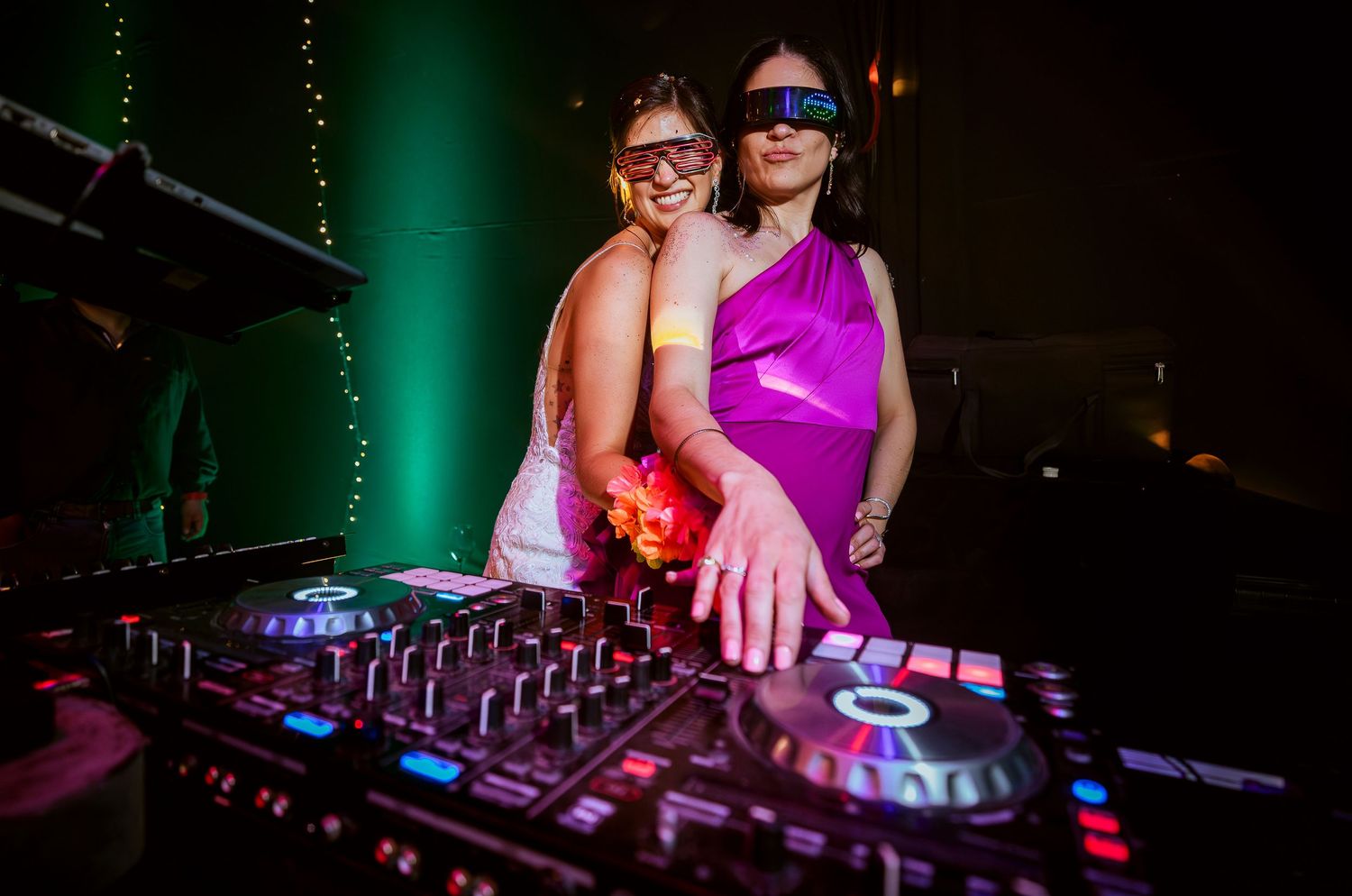 Two women wearing futuristic sunglasses pose together at a DJ mixer console at a vibrant nightclub event.