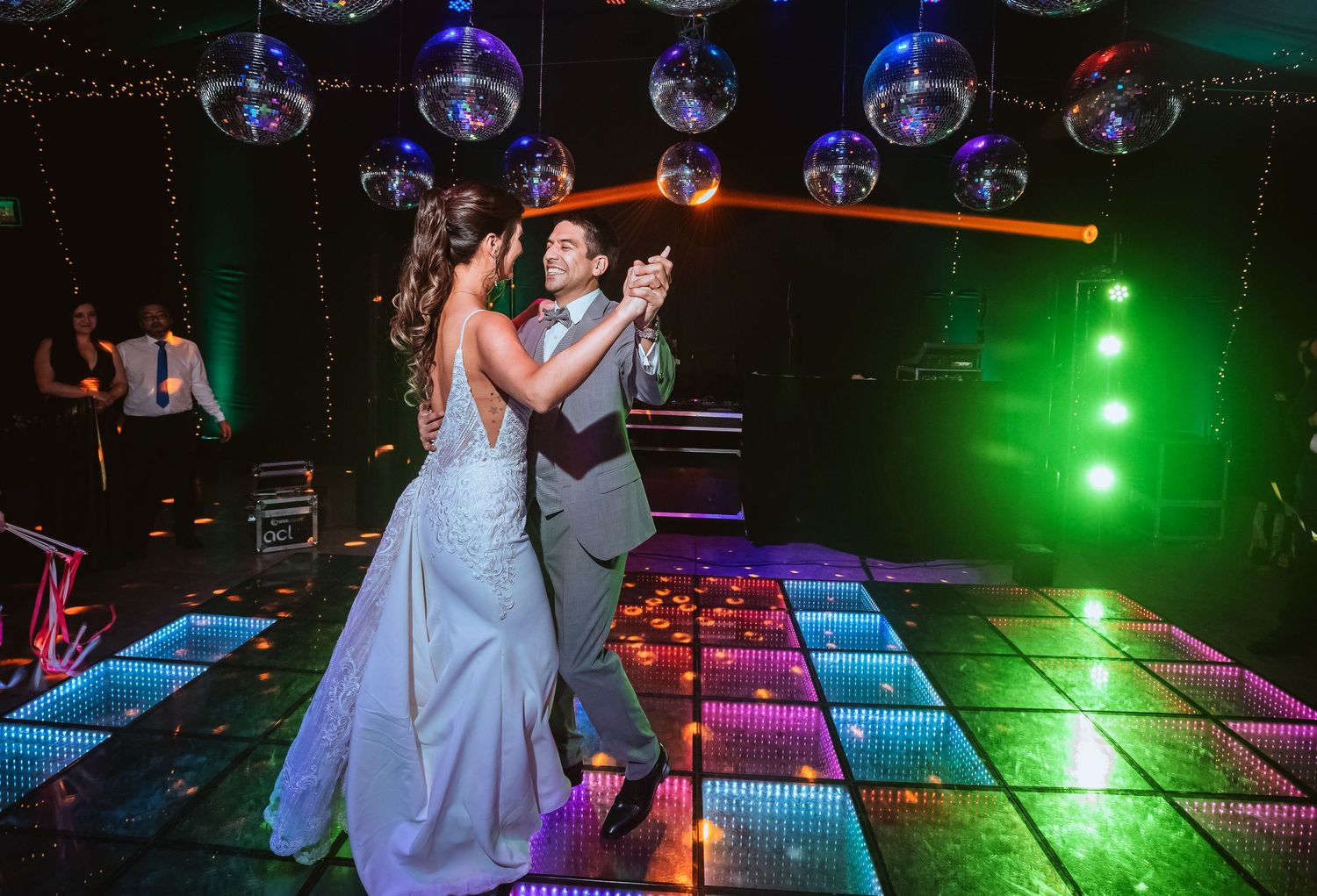 Couple sharing first dance on colorful LED dance floor with disco balls at elegant wedding reception.
