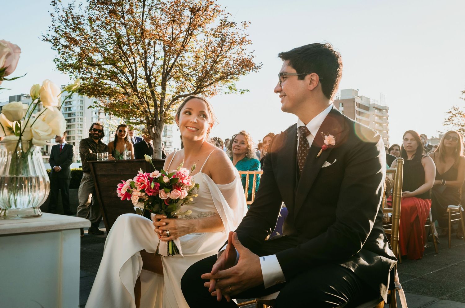 Bride holding pink bouquet sits beside groom during outdoor wedding ceremony.