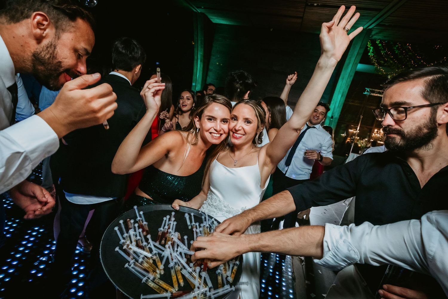 Bride and guests dance and celebrate around a tray of syringe shots at a wedding.