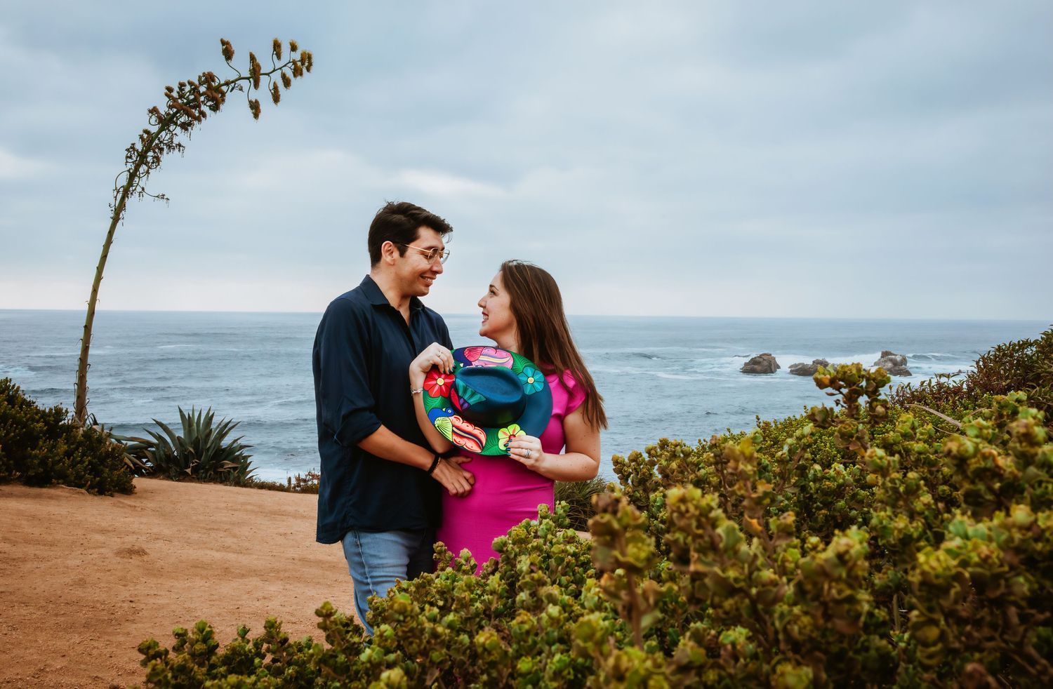 Couple smiling at each other holding colorful flowers near coastal cliffs with ocean views on a cloudy day.