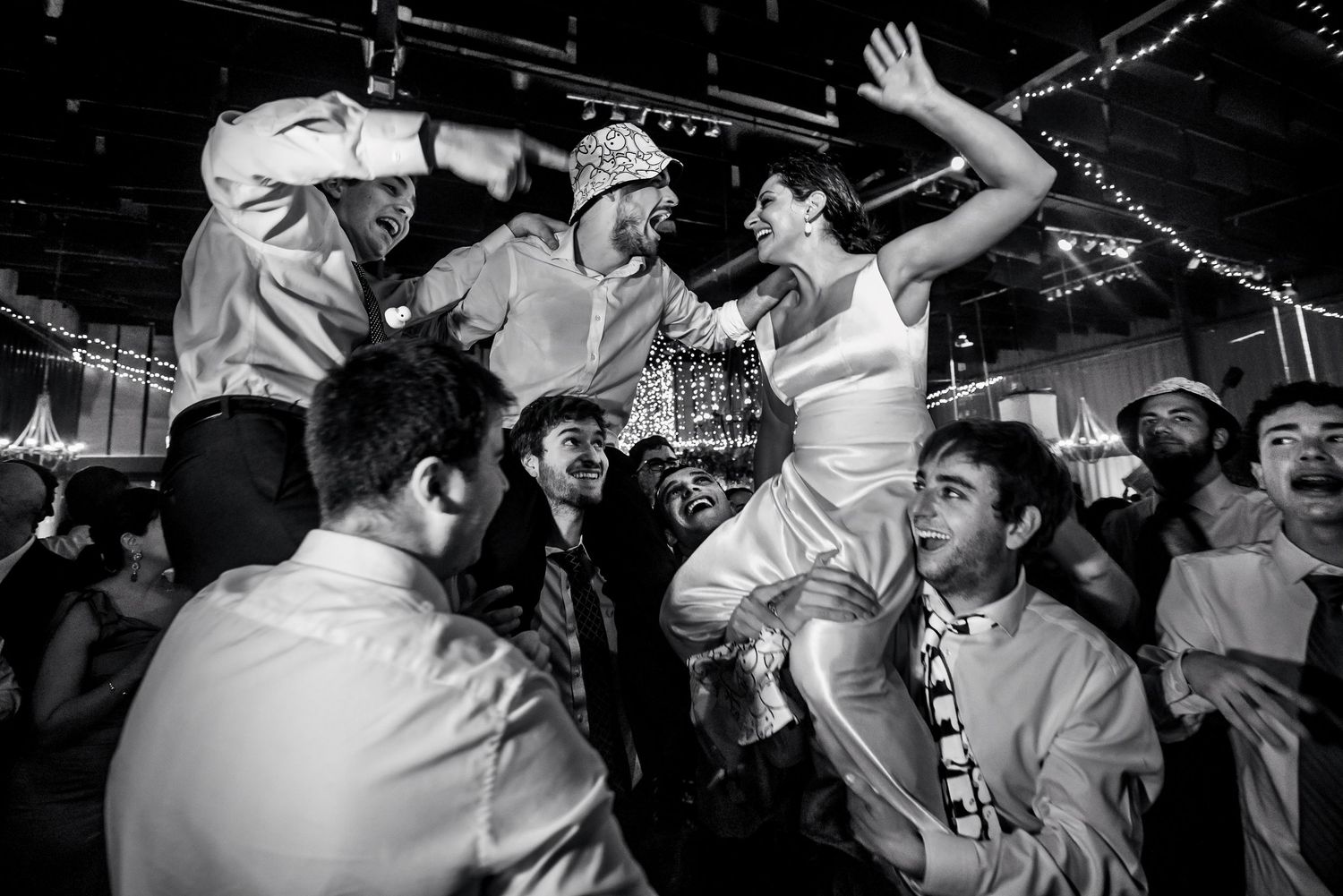 Black and white photo of bride and groom being lifted by guests during lively wedding reception dance celebration.