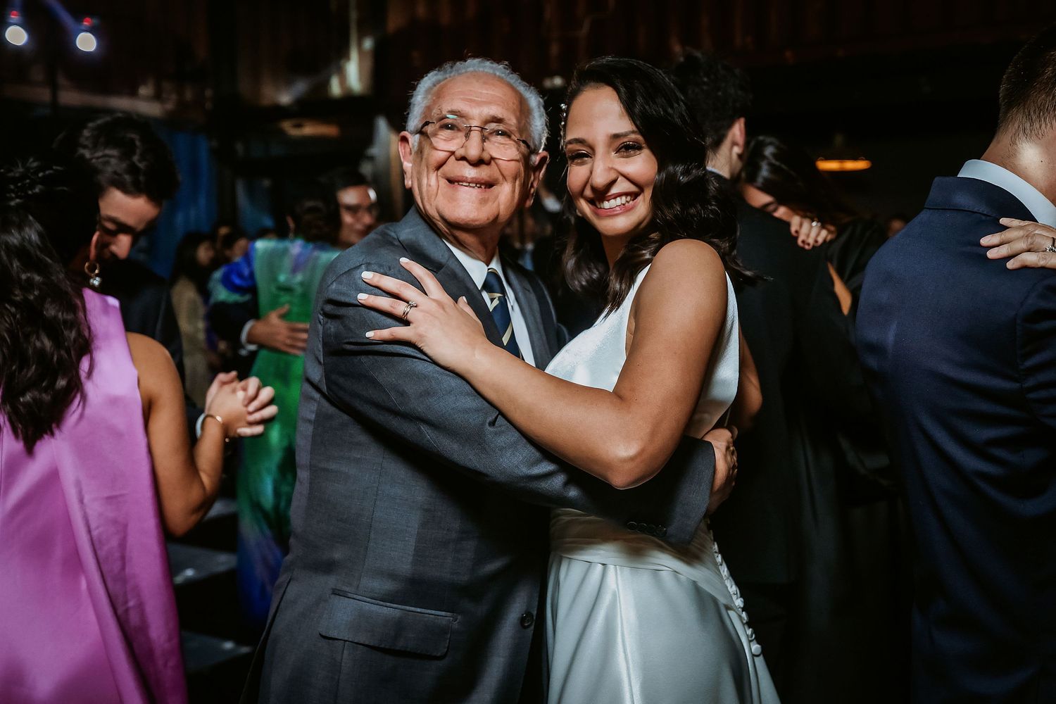 Bride in white dress hugs elderly man on wedding dance floor surrounded by guests.