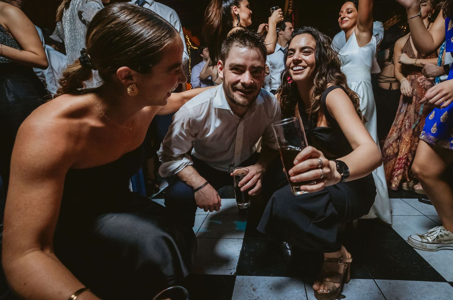 Three friends laughing and crouching on a dance floor at a party, holding drinks in a festive venue.