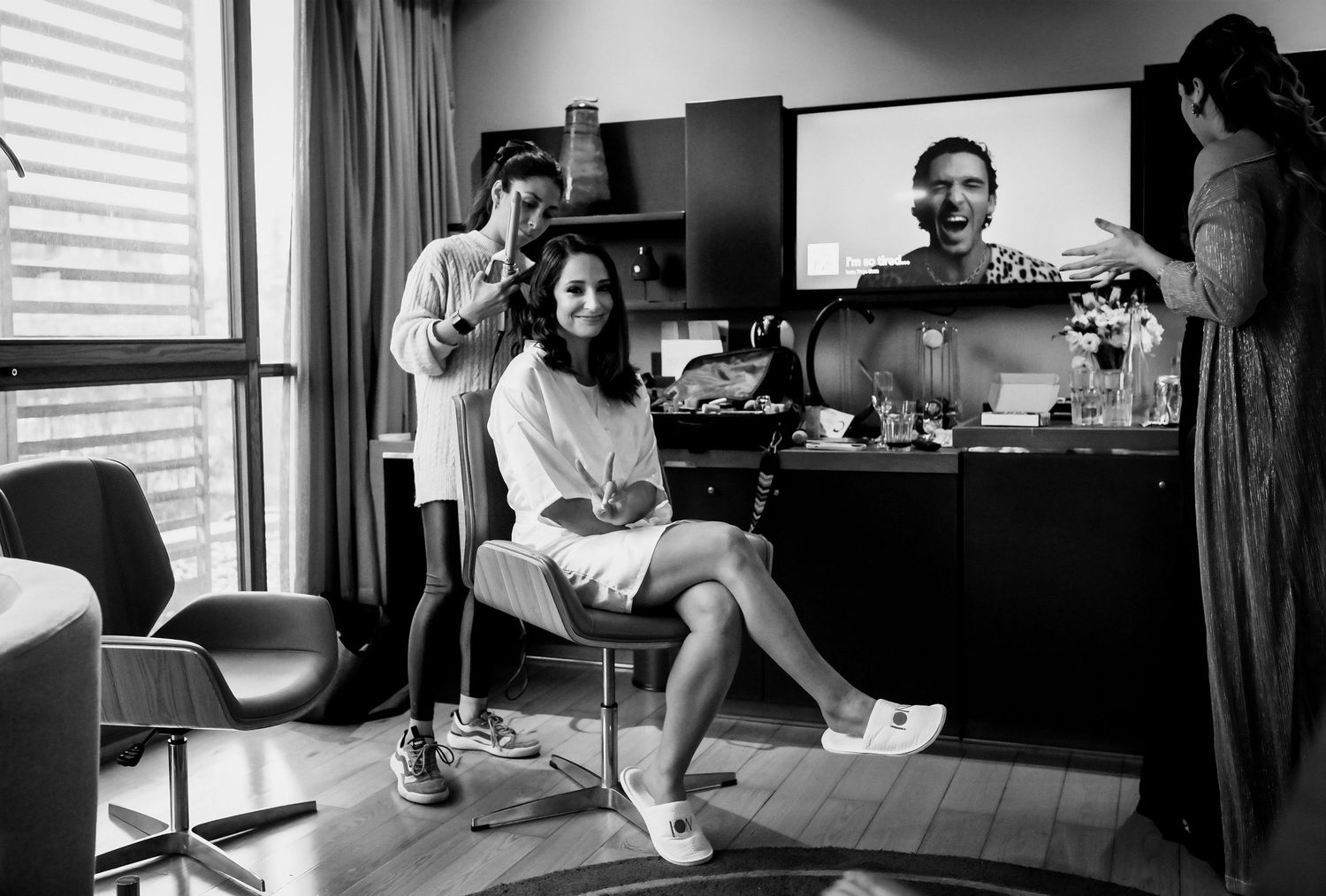 Black and white photo of a woman getting her hair done in a hotel room, smiling at the camera.