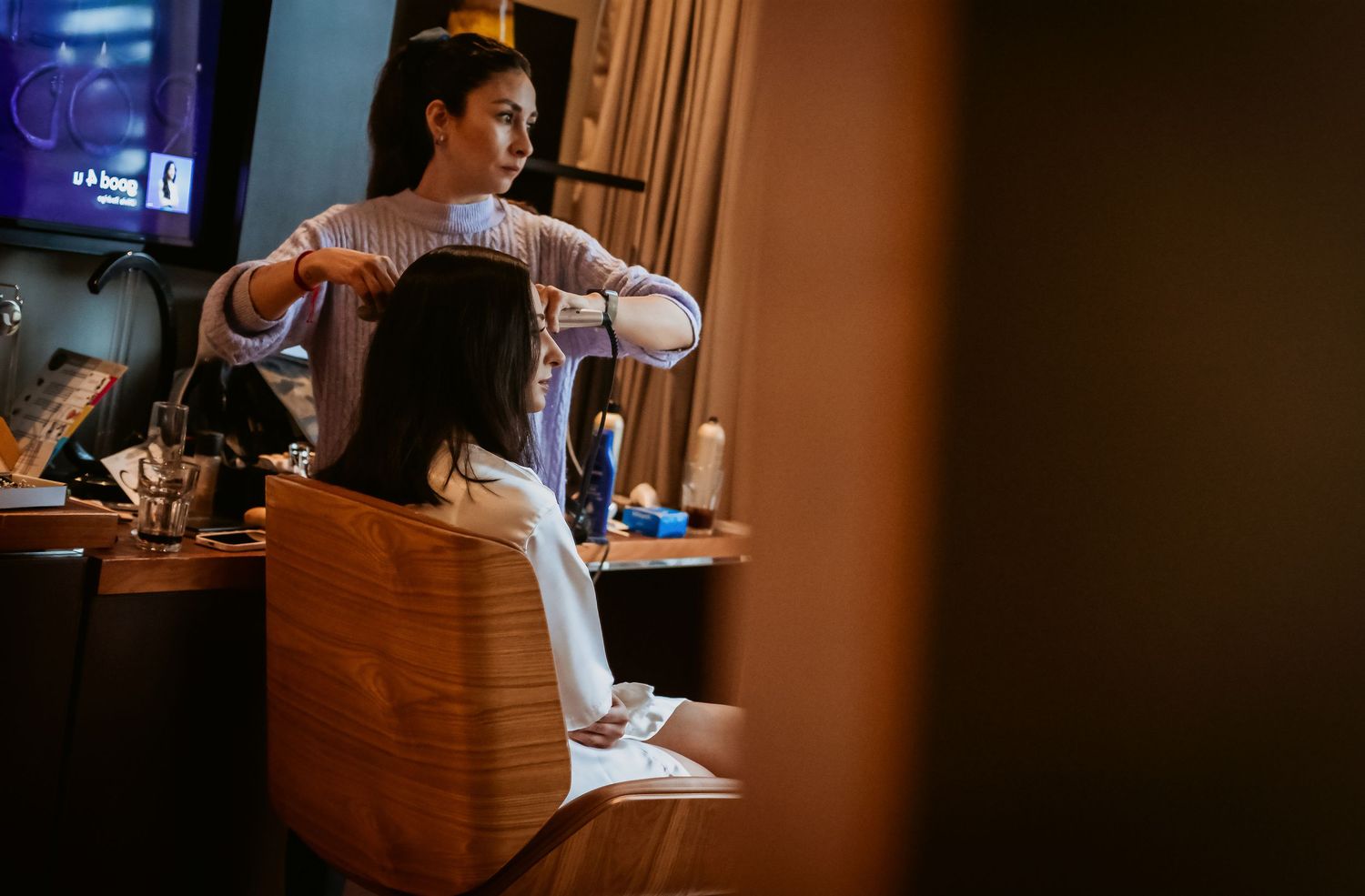 A hairstylist styling a seated woman's hair in a warmly lit salon with a mirror and TV in background.