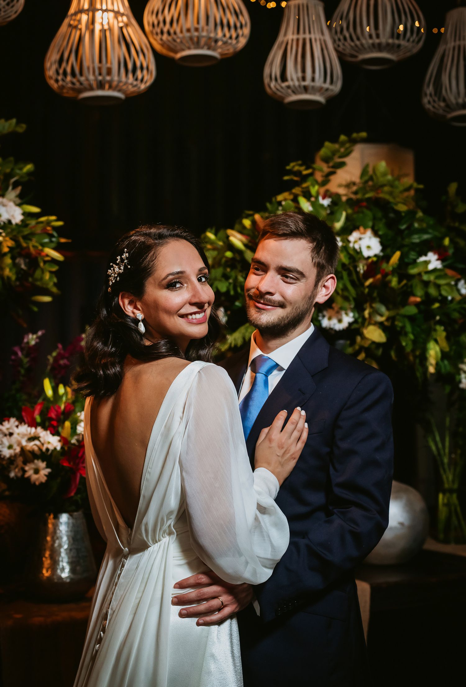 Bride in backless white gown and groom in navy suit with blue tie pose together at floral wedding reception.