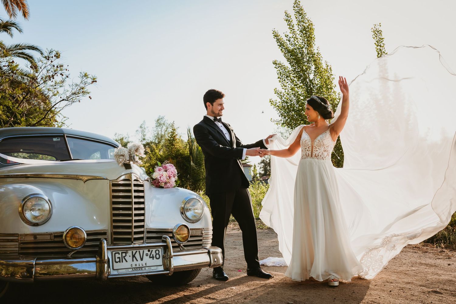Bride and groom pose beside a vintage classic car outdoors, with her white gown flowing in the breeze.
