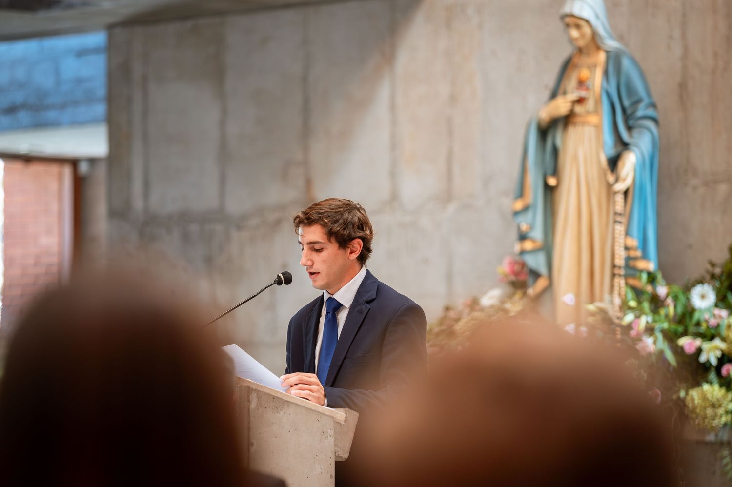 Young man in suit reads a passage at a church podium beside a statue of the Virgin Mary.