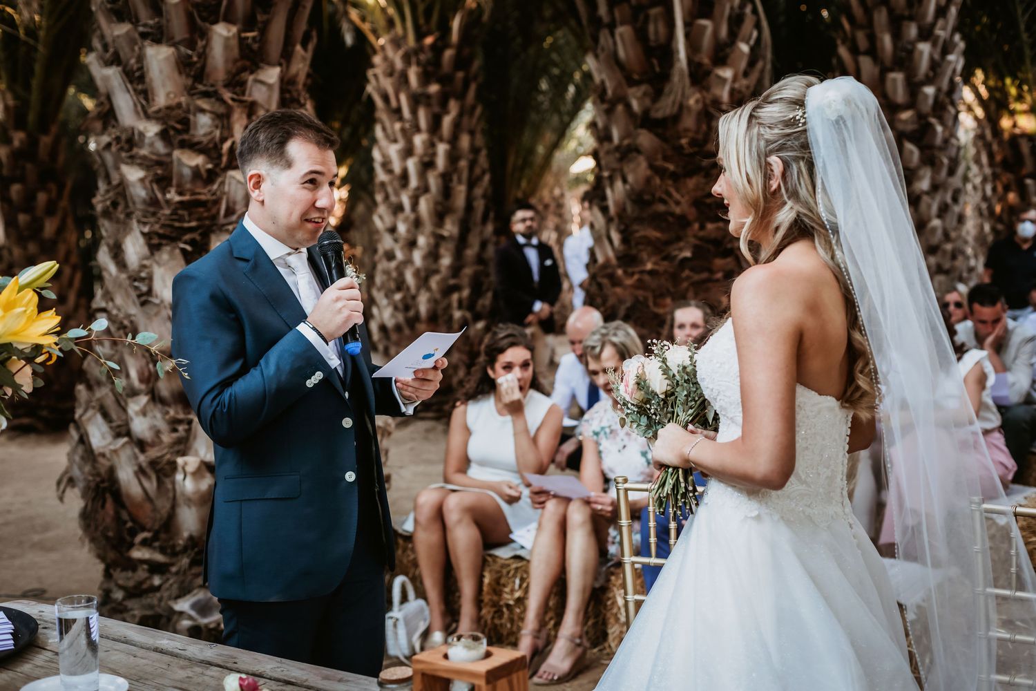 Groom in navy suit reads wedding vows to his bride at an outdoor ceremony beneath palm trees.