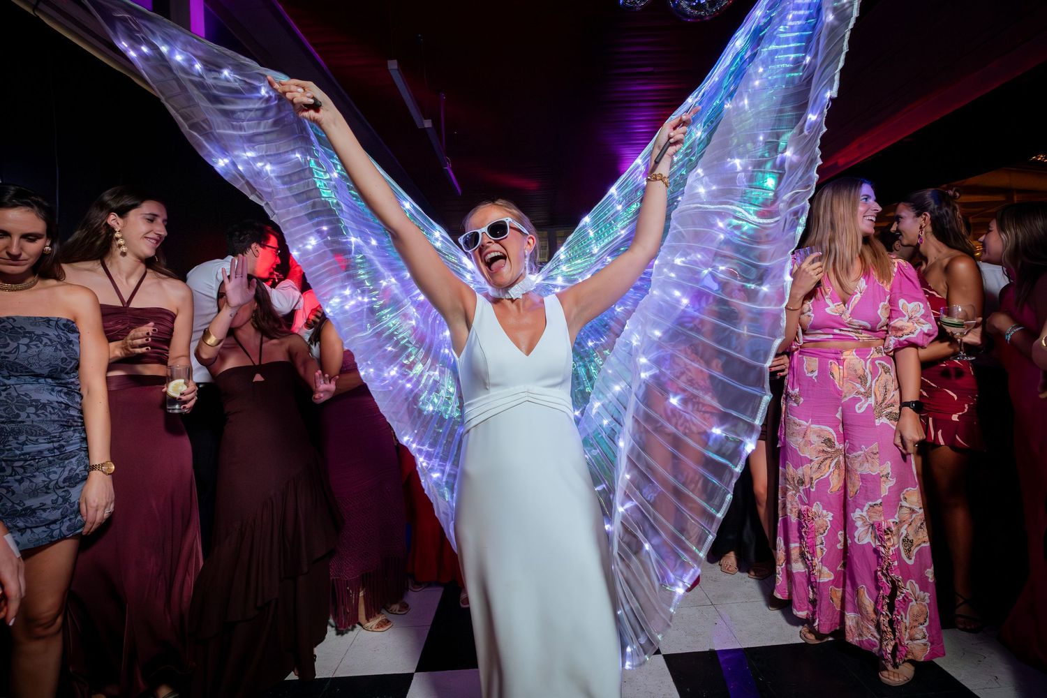 Bride in white dress wearing LED light-up angel wings and sunglasses dancing at wedding reception.