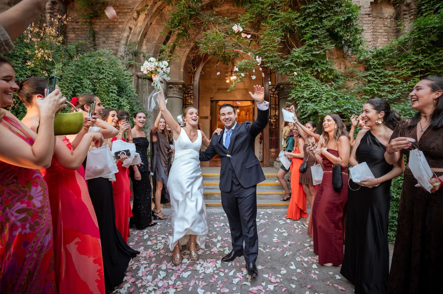 Bride and groom walk through confetti shower as wedding guests celebrate in elegant courtyard venue.