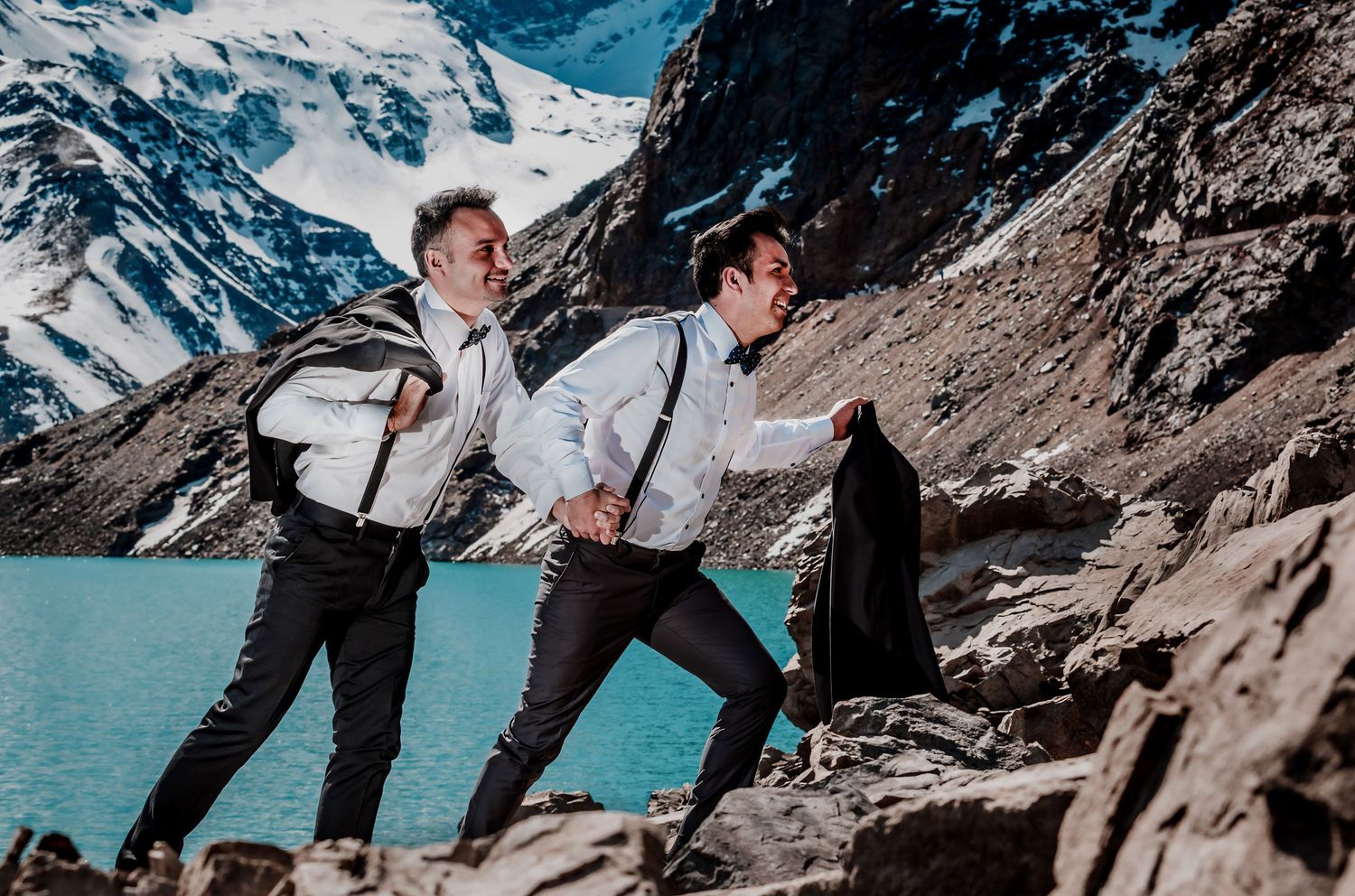Two grooms in suspenders walk playfully across rocky terrain by a bright turquoise glacial lake.