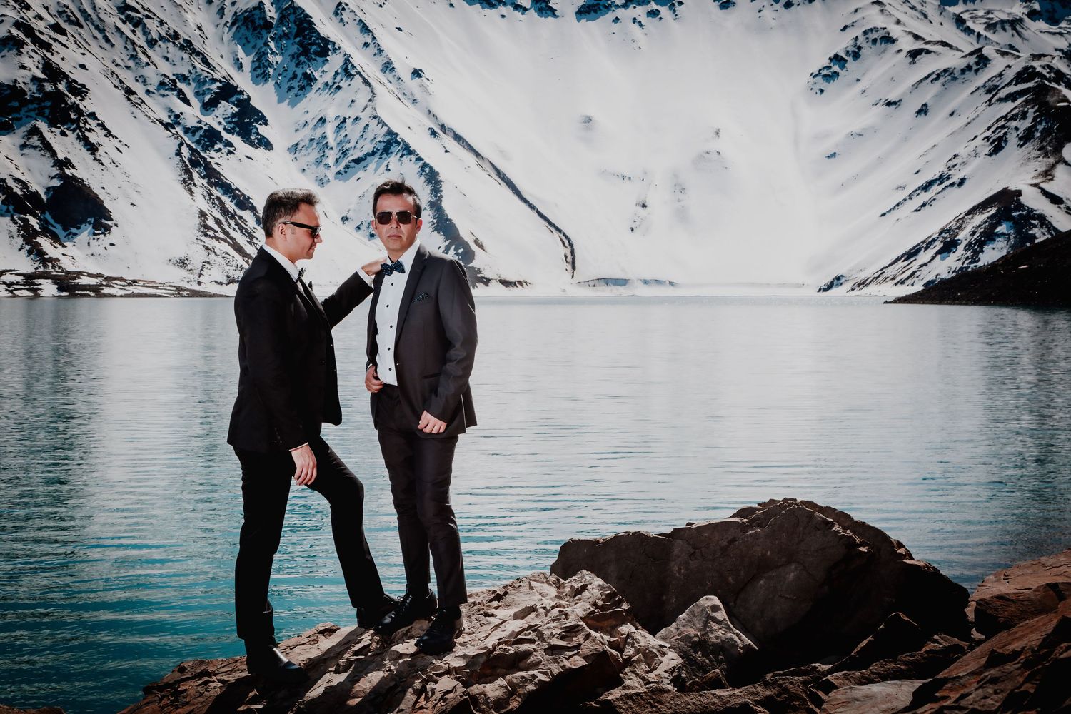 Two men in suits share a playful moment on rocks beside a vivid blue glacial mountain lake.