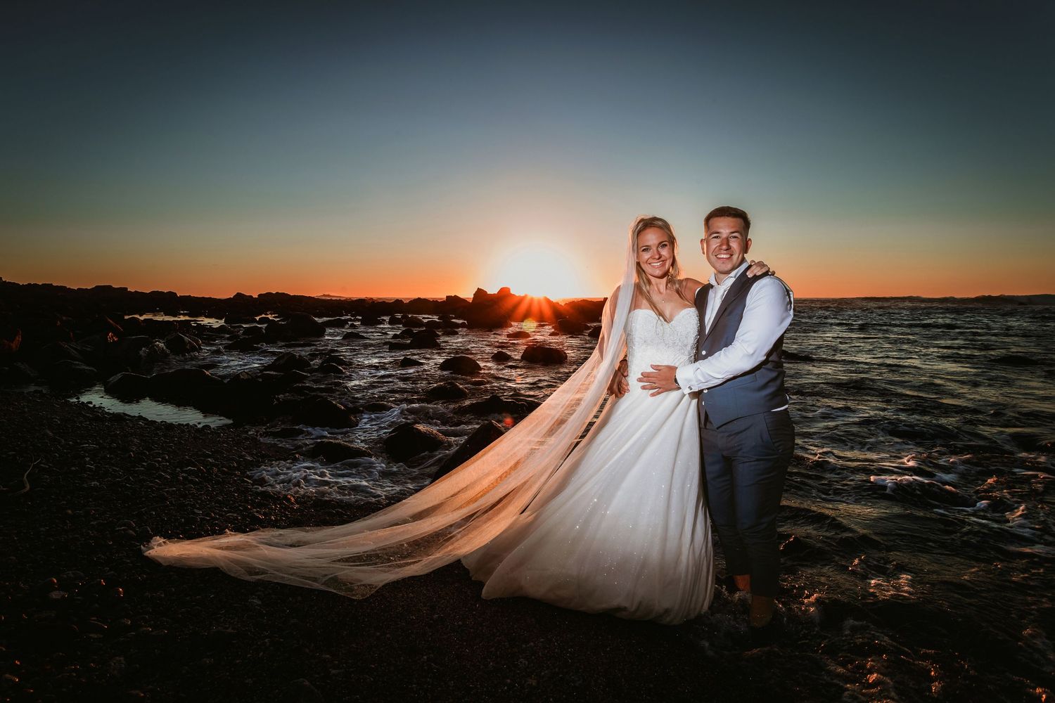 Bride and groom embracing on rocky shoreline with stunning golden sunset behind them.