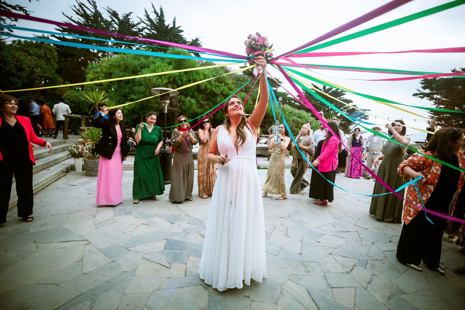 Bride raises colorful ribbon bouquet high during festive outdoor wedding bouquet toss ceremony.