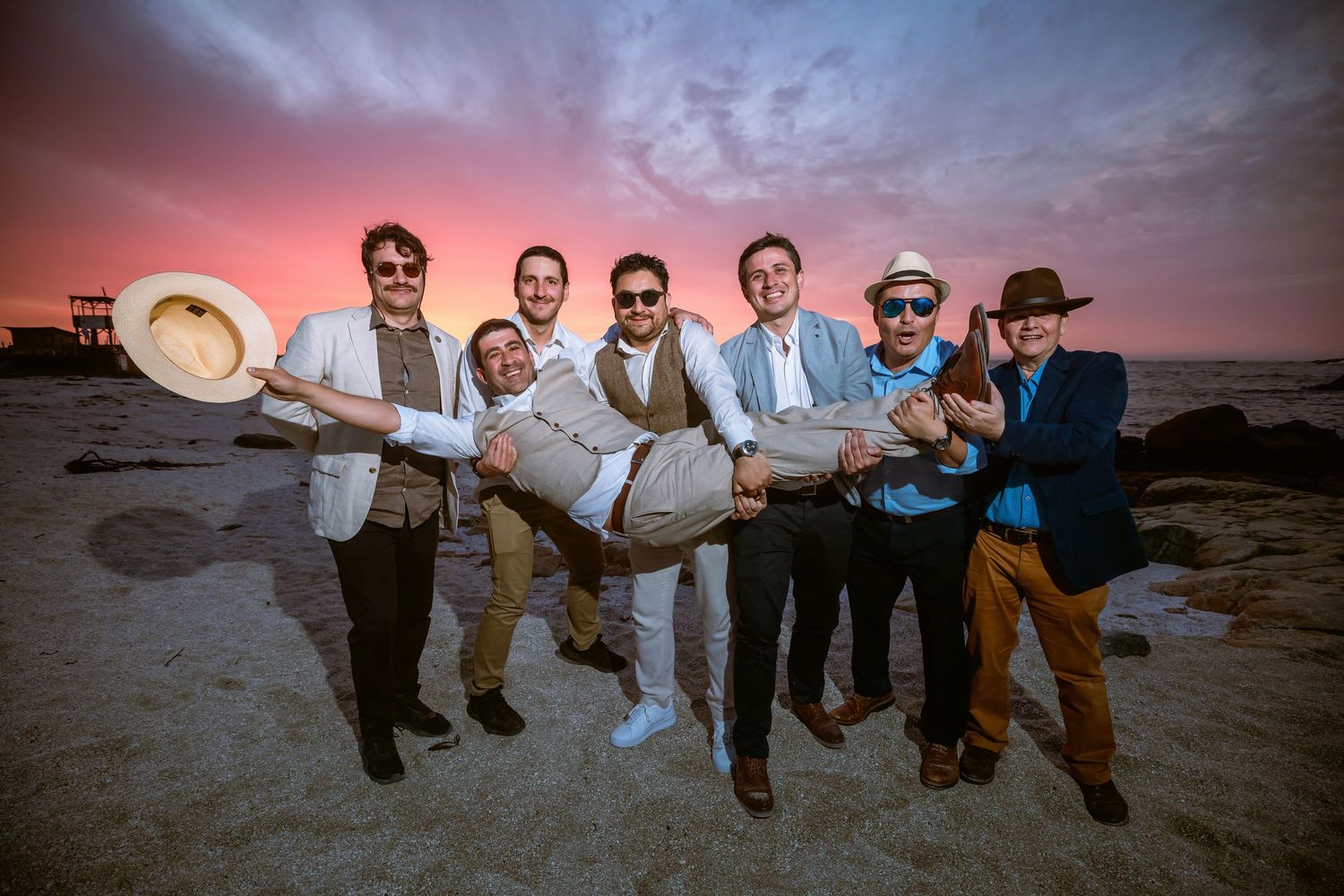Groomsmen lifting groom on beach during colorful pink and purple sunset.