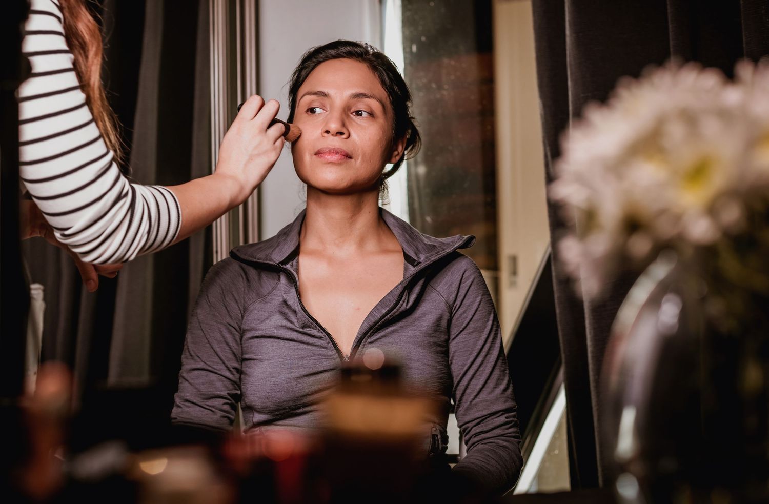 A makeup artist applies foundation to a woman seated near a floral arrangement.