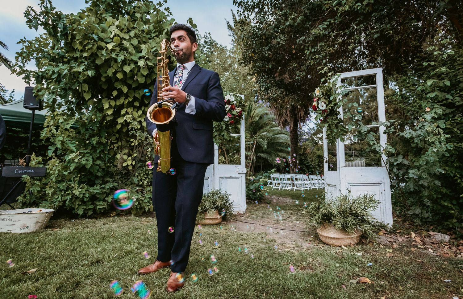 Elegant saxophonist in suit performing outdoors at garden wedding surrounded by confetti and greenery.