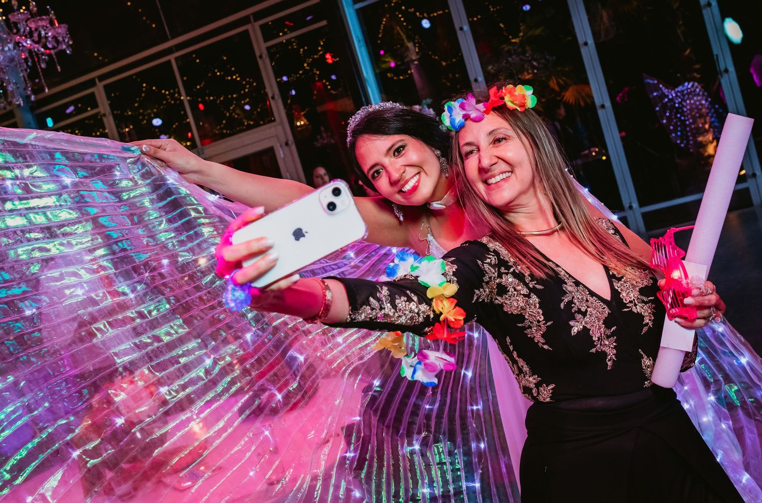 Two women taking a selfie at a wedding party with glowing LED wings and floral crowns.