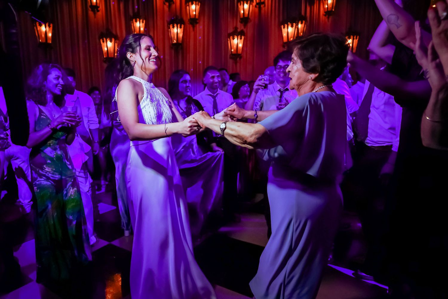 Two women in elegant gowns dance together under purple lights at a wedding reception party.