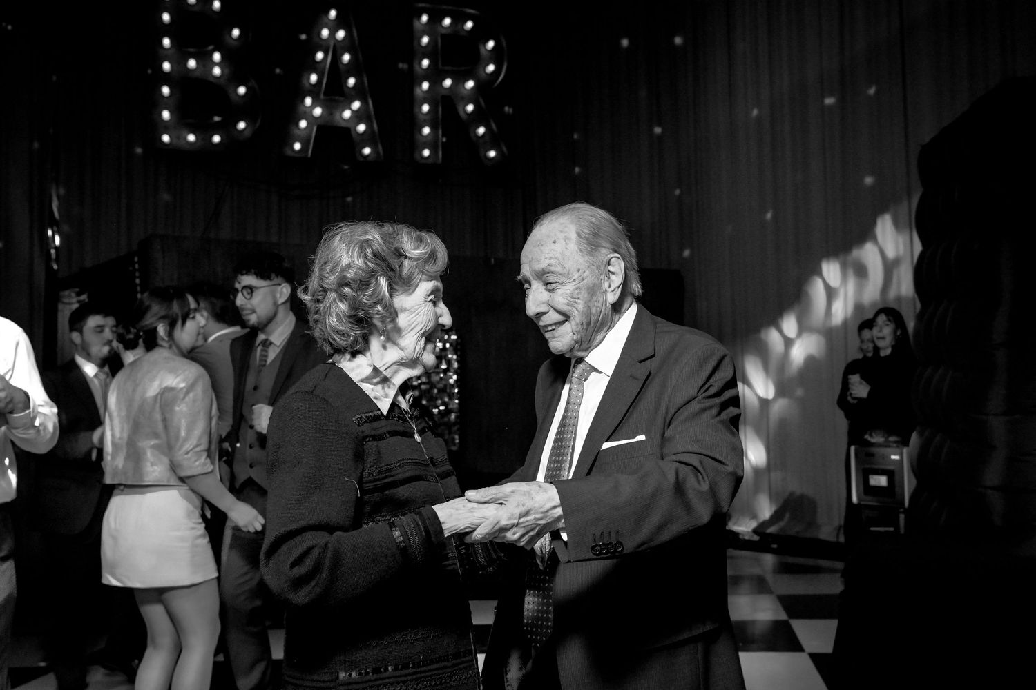 Elderly couple dancing together at wedding reception in black and white photo near illuminated BAR sign.