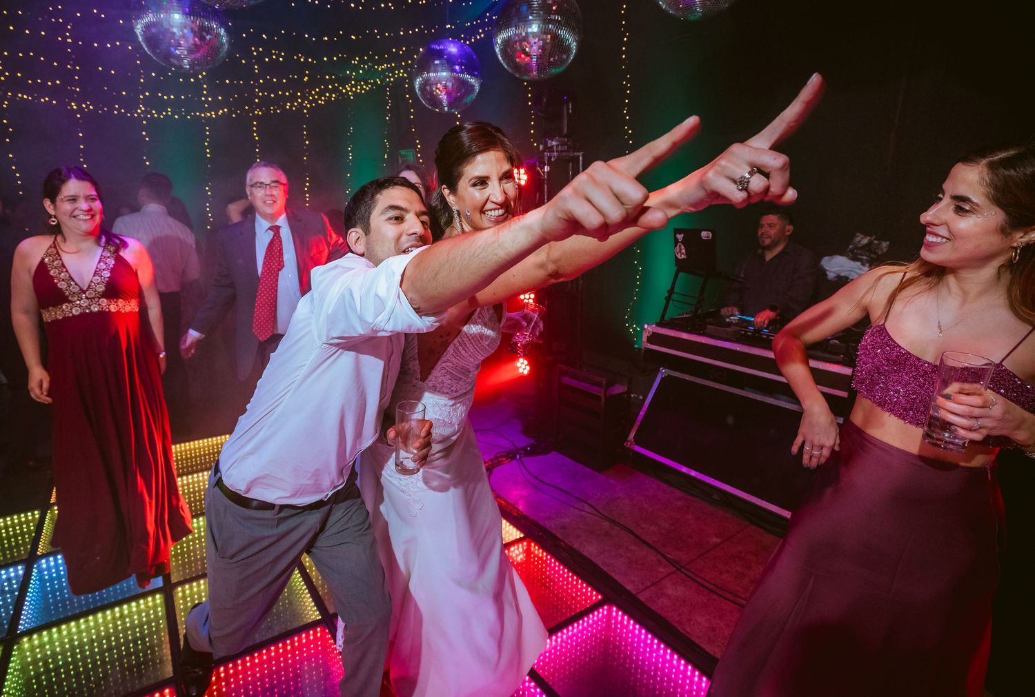 Couple dancing joyfully at a wedding reception on a colorful LED dance floor surrounded by guests in formal attire.