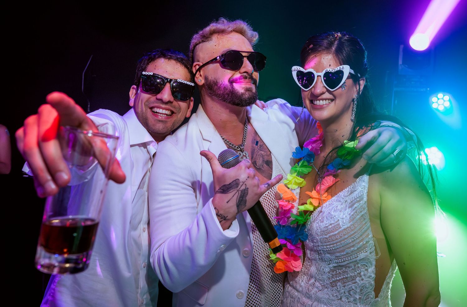 Three friends in white outfits pose at a neon-lit party, wearing fun accessories like heart sunglasses and a rainbow lei.