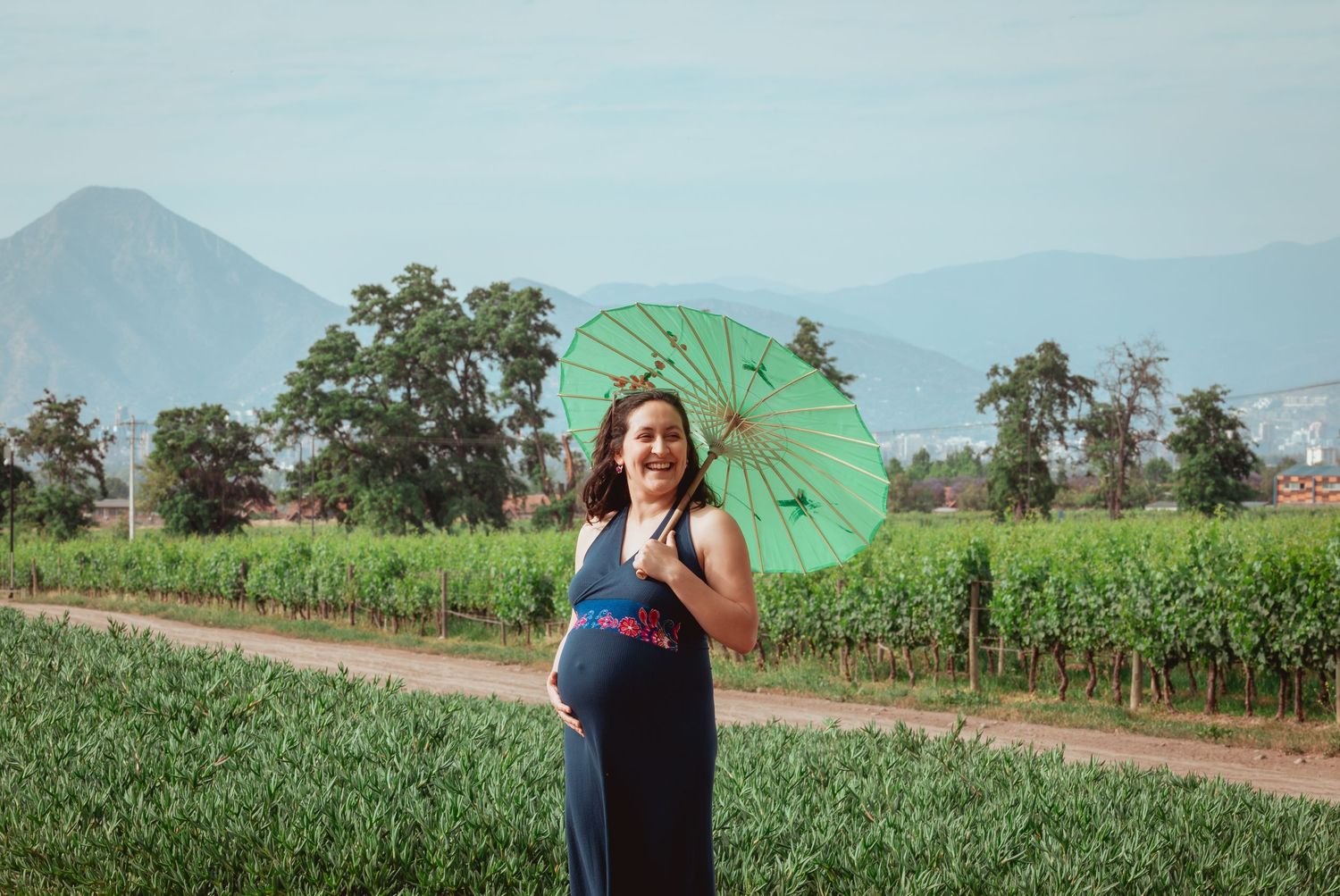 Pregnant woman smiling with green parasol in a sunny green farmland setting.