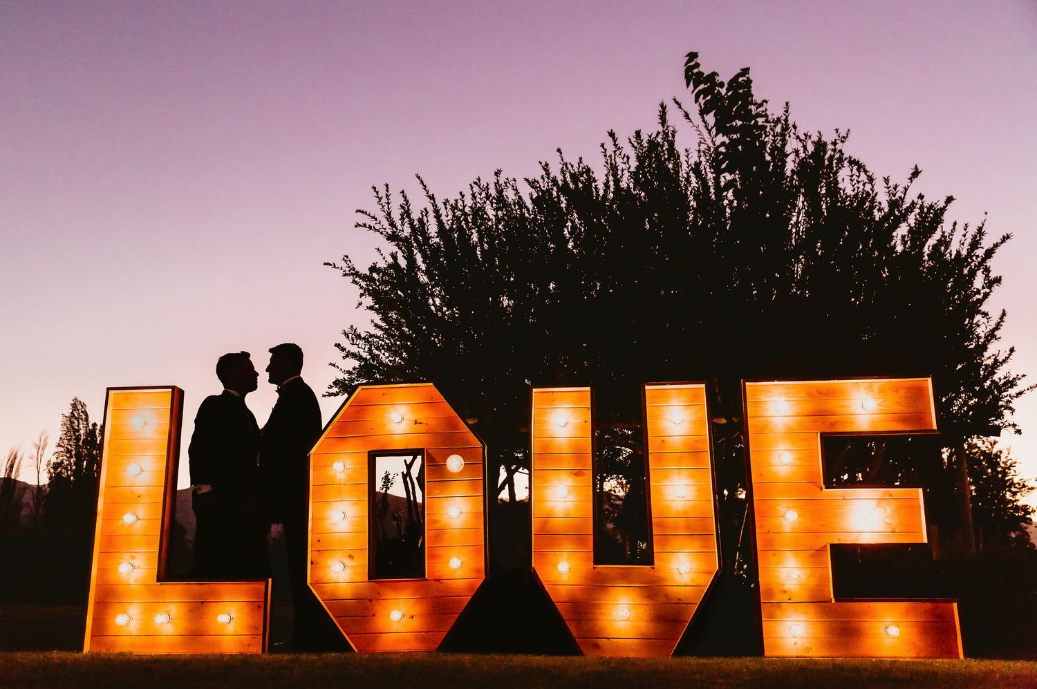 Couple silhouetted against twilight sky beside illuminated LOVE marquee letters at outdoor wedding celebration.