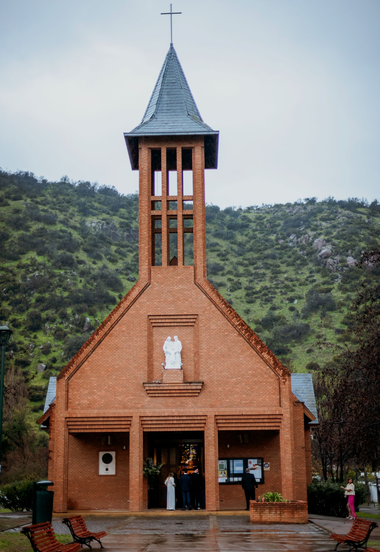 Red brick Catholic church with bell tower and cross, set against green hills on a cloudy day.