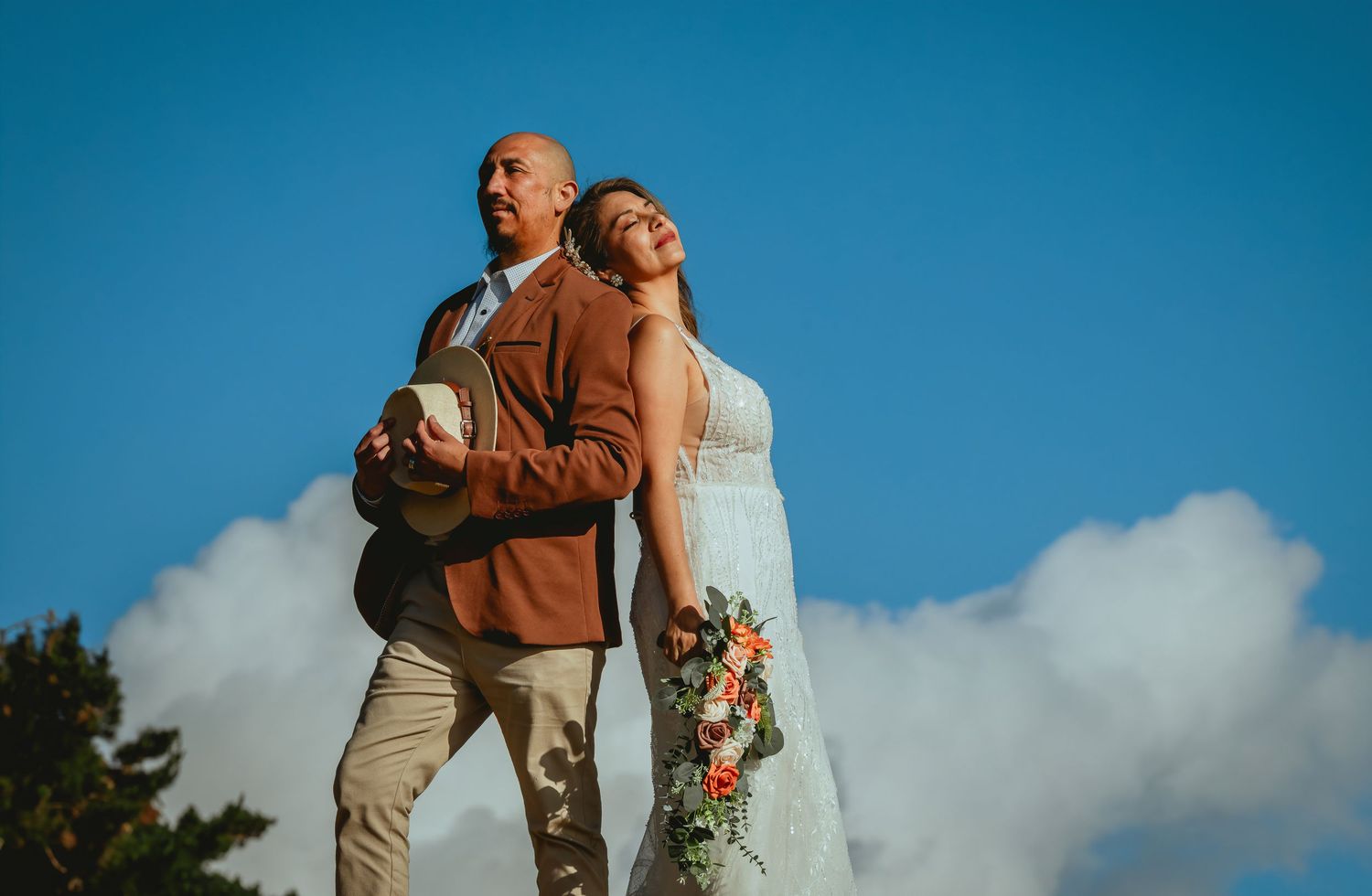 Bride in a white lace gown holding a floral bouquet stands back to back with her groom in a brown blazer against a blue sky and clouds.