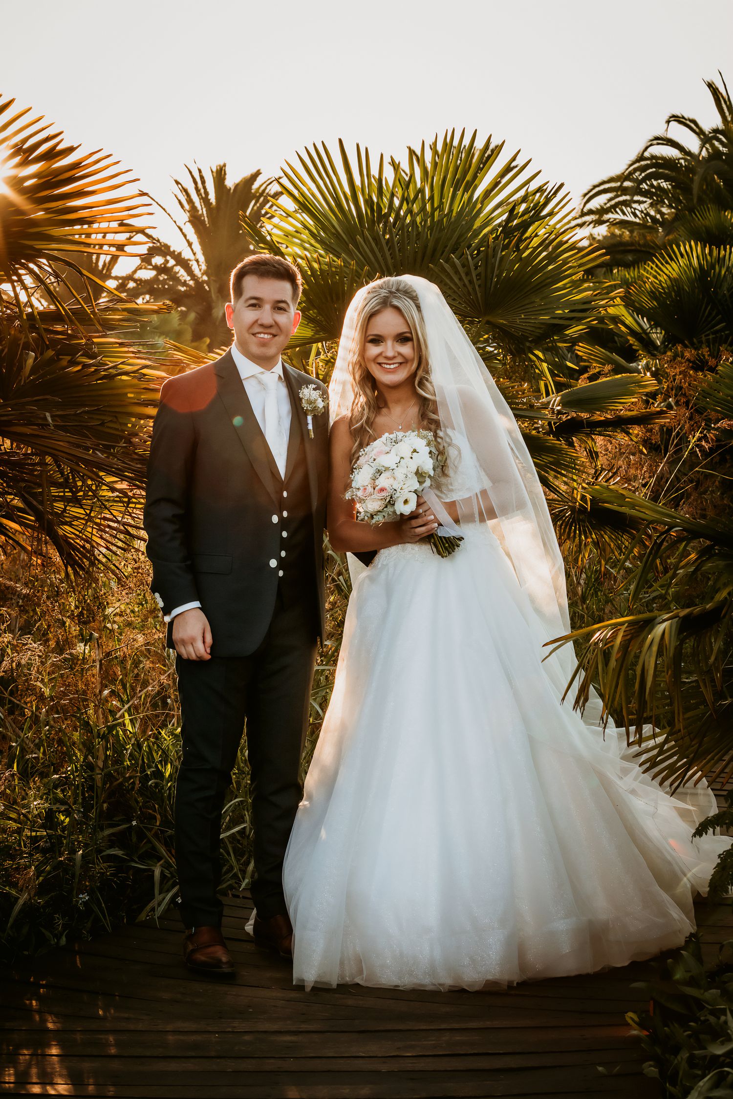 Bride and groom posing on a wooden boardwalk surrounded by tropical palms at sunset.
