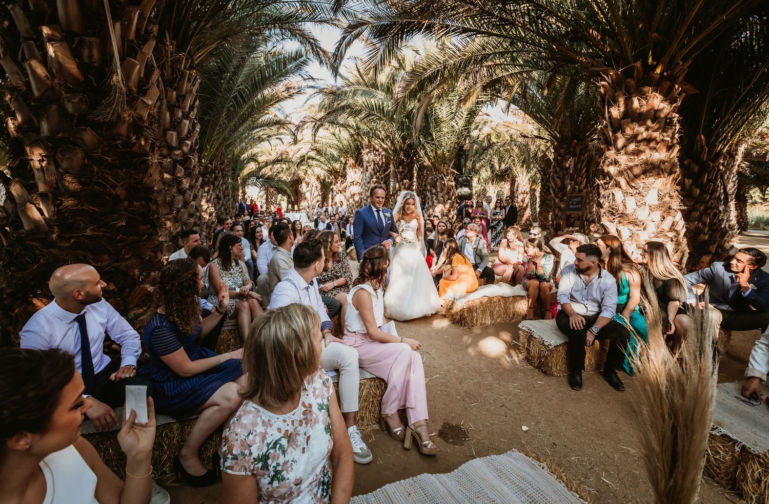 Bride and groom walking down outdoor wedding aisle surrounded by palm trees and guests.