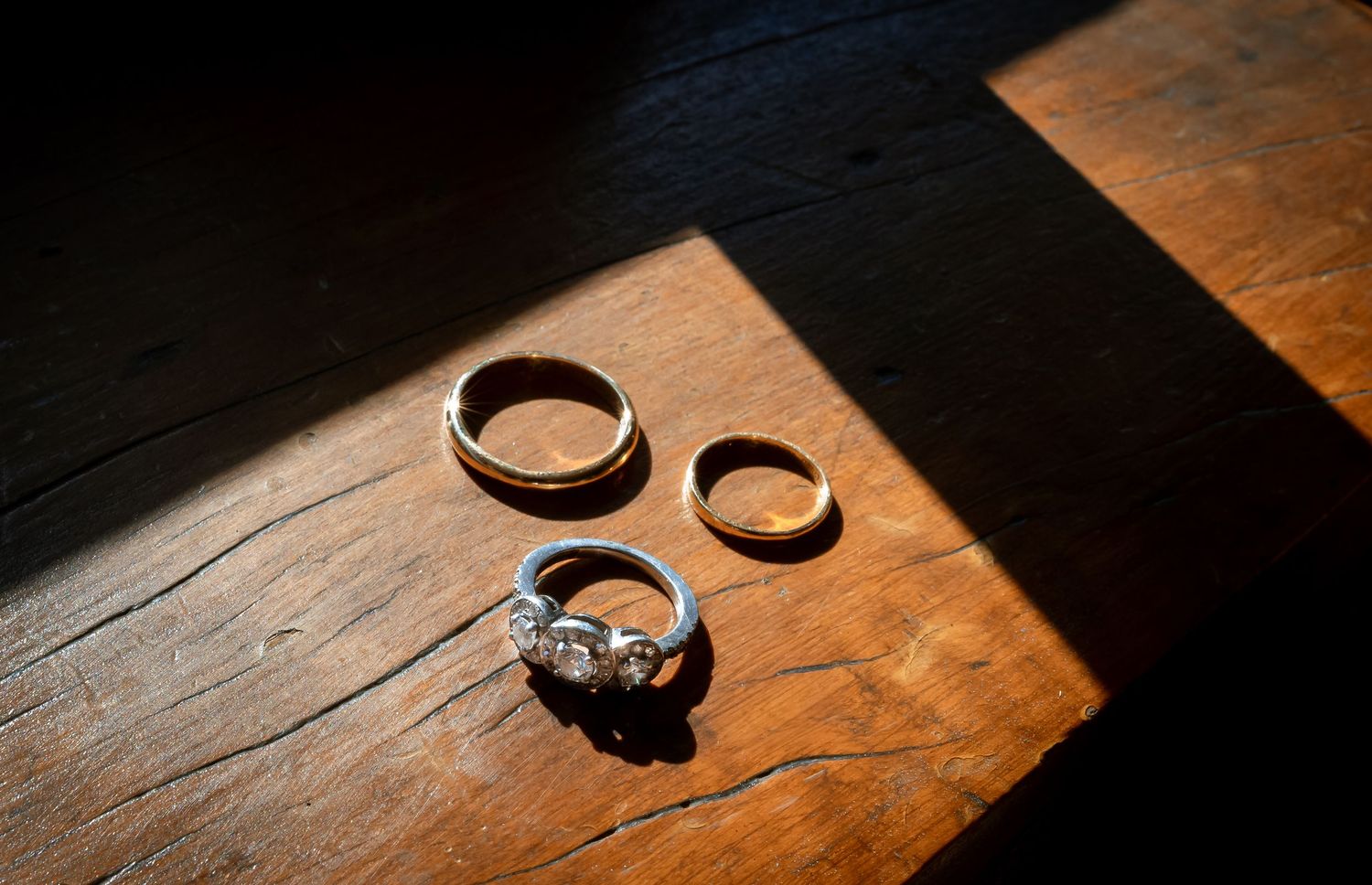 Three wedding rings, including a diamond engagement ring, on a rustic wooden floor in sunlight.