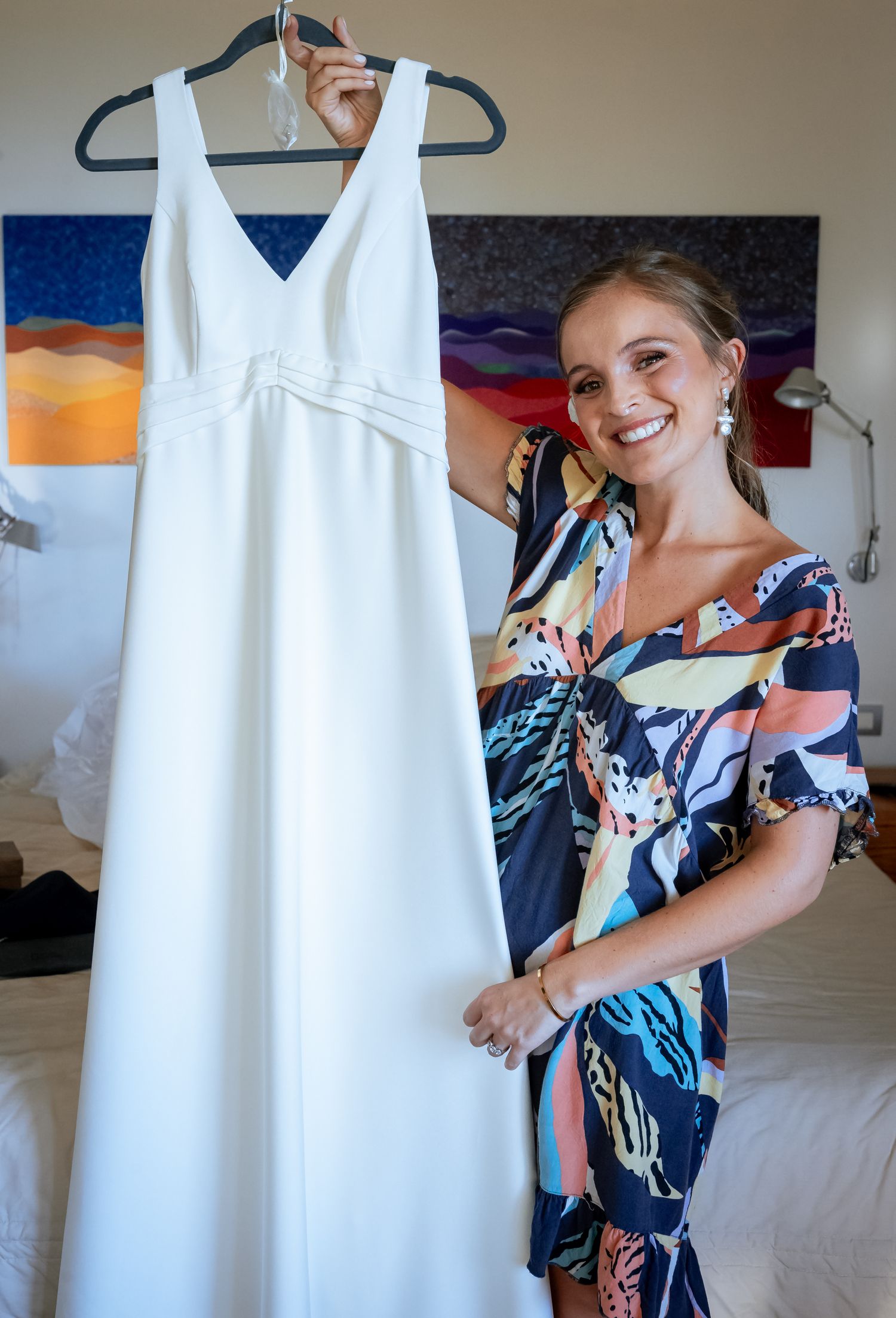 Bride holding white wedding gown on hanger in hotel room wearing floral robe.