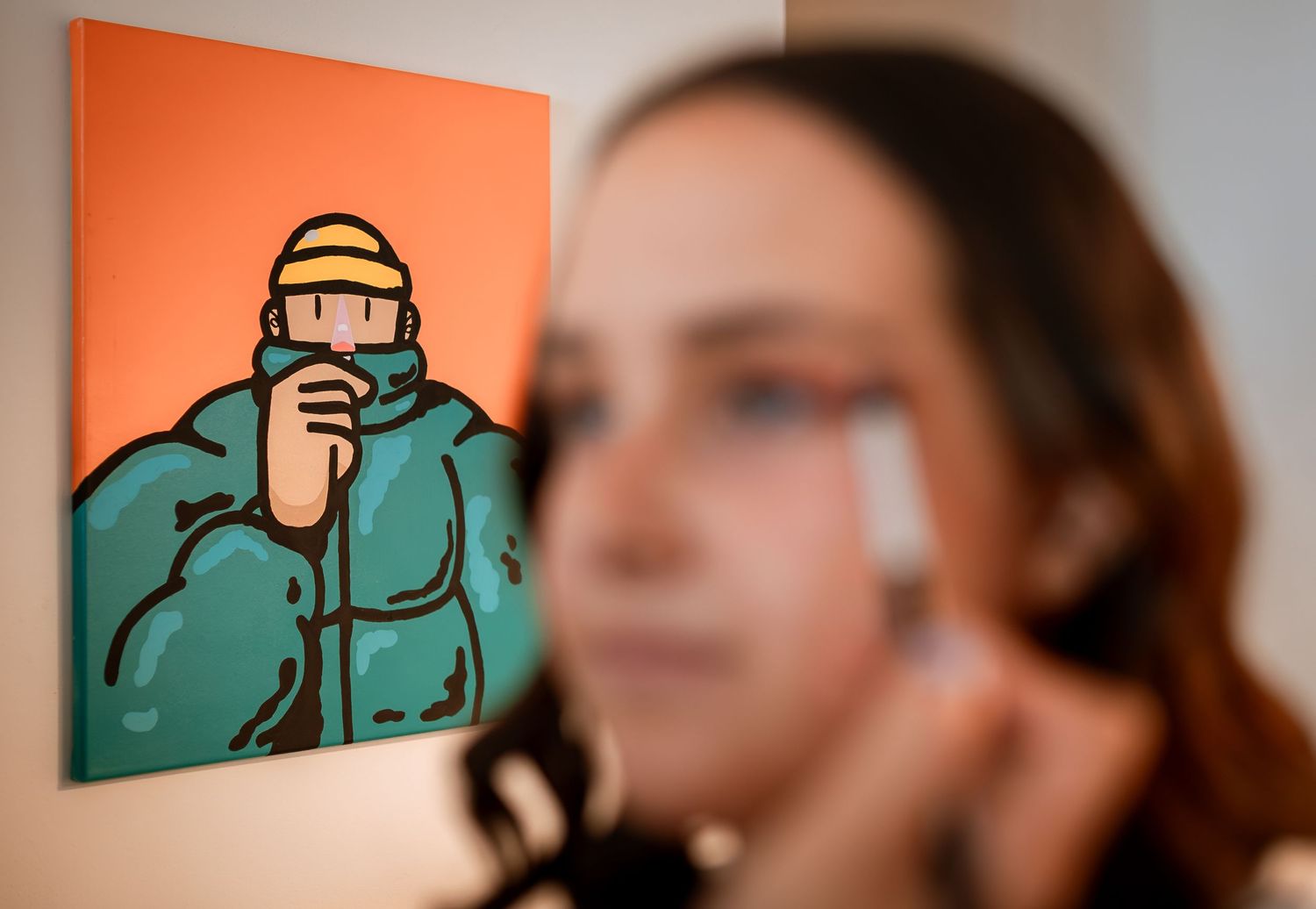 Woman applying makeup in foreground with a colorful pop art painting visible in the background.