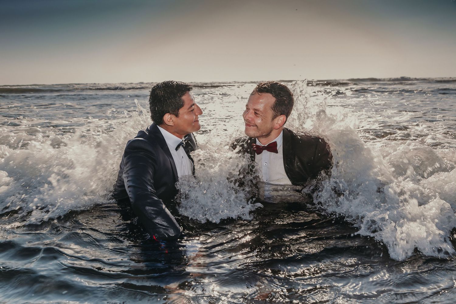 Two grooms in tuxedos laugh together as waves crash around them during a beach wedding photo session.