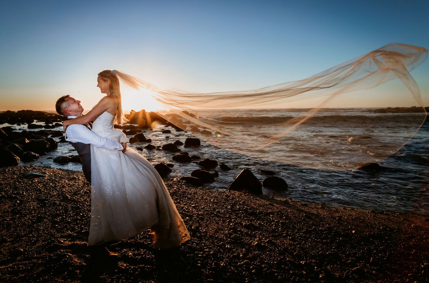 Groom lifting bride on rocky beach at sunset, her veil flowing dramatically in the wind.
