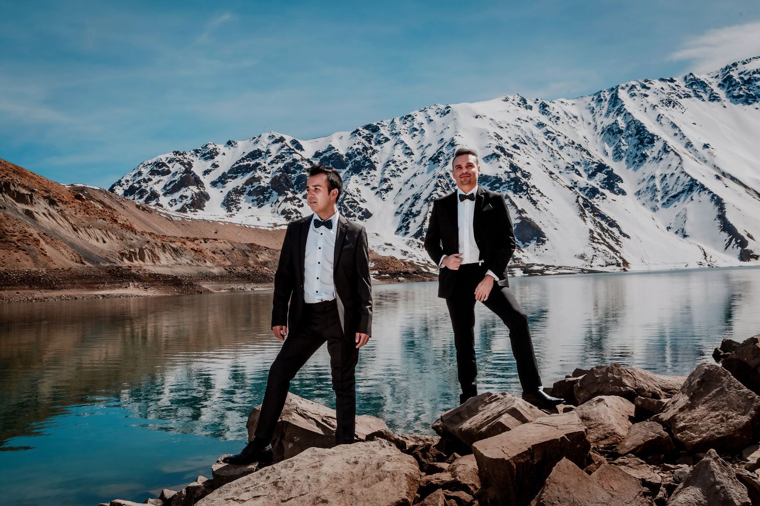Two grooms in tuxedos pose on rocks beside a glacial lake with snowy mountains behind them.