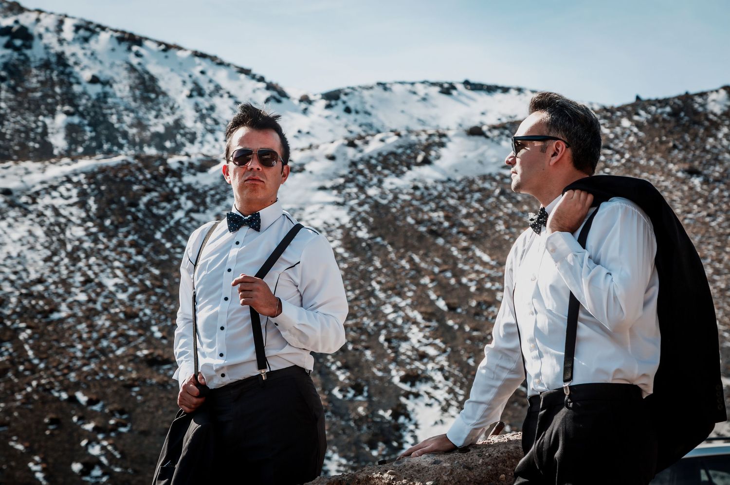 Two grooms wearing sunglasses and suspenders pose against a dramatic snow-dusted mountain backdrop.