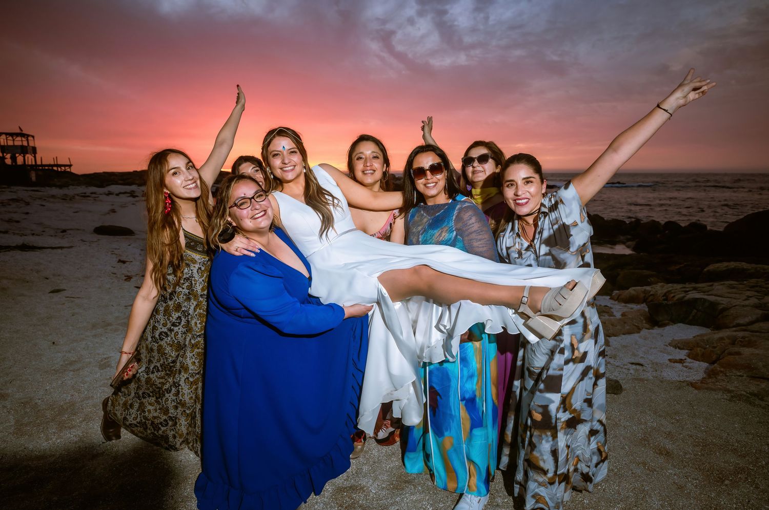 Bridesmaids joyfully lifting bride on sandy beach during vibrant pink sunset.